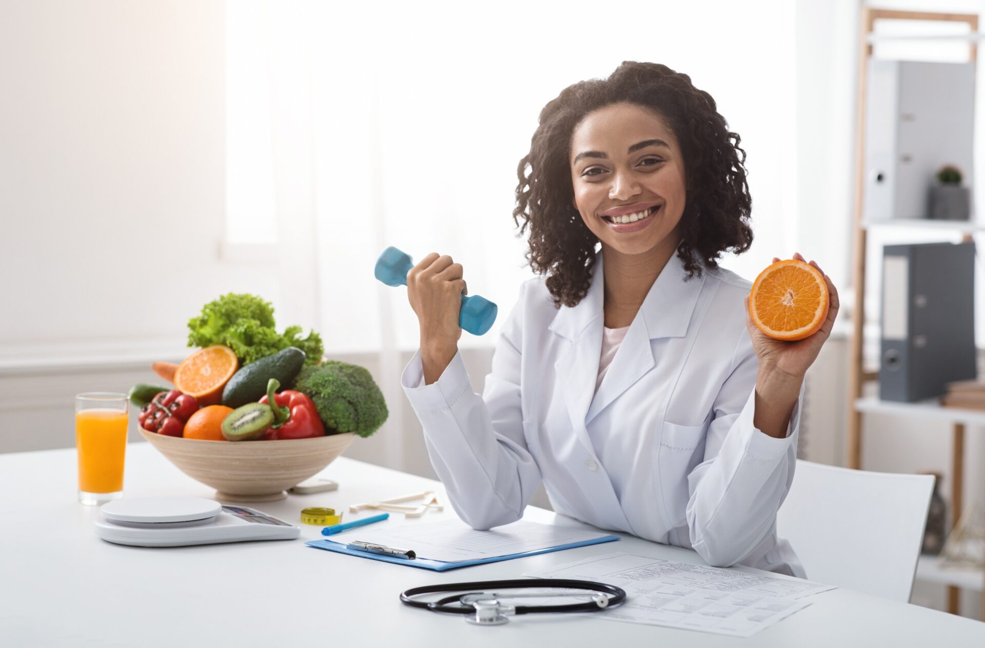 Sport and vitamins for immunity. Happy black woman doctor holding orange and barbell in clinic, posing at her workplace, free space Sport and vitamins for immunity. Happy black woman doctor holding orange and barbell in clinic, posing at her workplace, free space